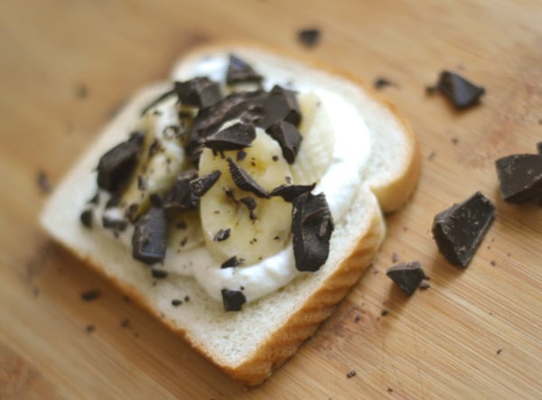 A slice of white bread topped with yogurt, banana slices, and chunks of dark chocolate creates a delightful twist on a banana and chocolate toasted sandwich, served on a rustic wooden surface.