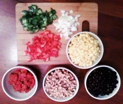 A wooden cutting board with chopped green bell pepper, onion, and tomato, next to bowls of corn, diced tomatoes, black-eyed peas, and black beans on a wooden table.
