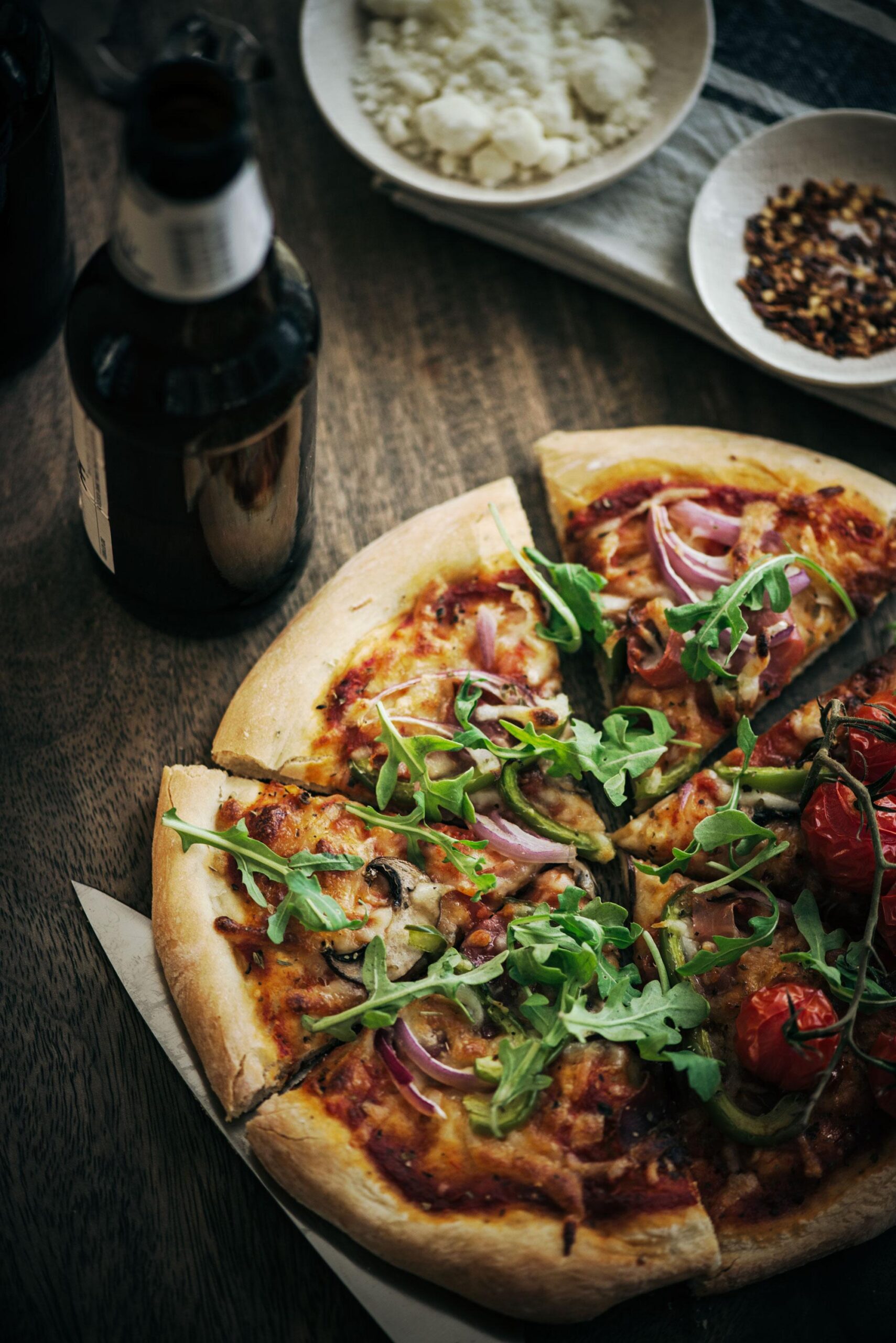 A sliced homemade pizza topped with arugula, red onions, and roasted tomatoes sits on a wooden table next to a bottle of beer, a bowl of crumbled cheese, and a dish of red pepper flakes.