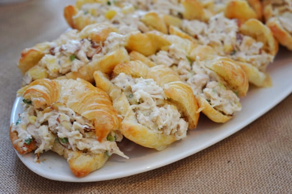 Mini croissants filled with creamy chicken salad, arranged closely on a white serving platter placed on a burlap tablecloth.
