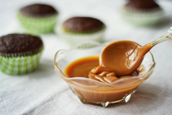 A glass bowl filled with rich caramel sauce, with a spoon lifting some out. In the blurred background, four chocolate cupcakes in green checkered paper liners sit on a white surface.