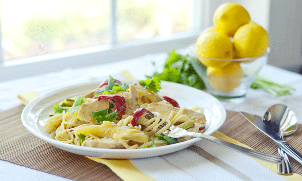 A plate of chicken linguine with lemon sauce, red bell peppers, and greens sits on a table next to a fork and spoon. In the background, a glass bowl of lemons rests near a sunlit window.