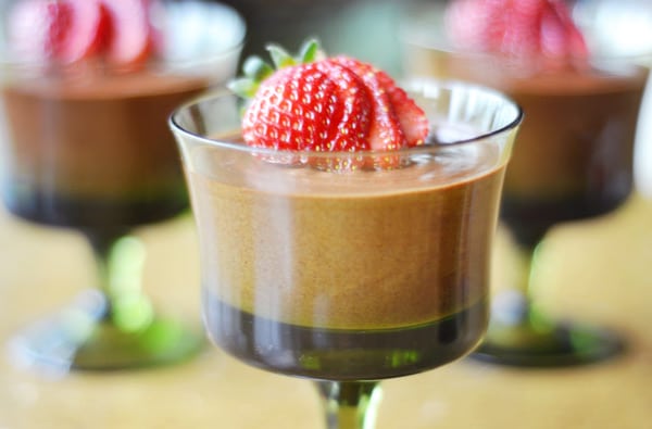 A glass dessert cup filled with layers of rich chocolate mousse, topped with a fresh, sliced strawberry. Two similar chocolate mousse dessert cups are blurred in the background.