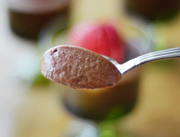 A close-up of a spoonful of rich chocolate mousse, with a blurred background showing a glass dessert topped with a red strawberry.