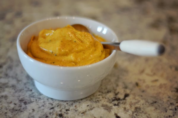 A small white bowl filled with creamy orange sauce or dip, with a white-handled spoon resting inside, placed on a speckled countertop.