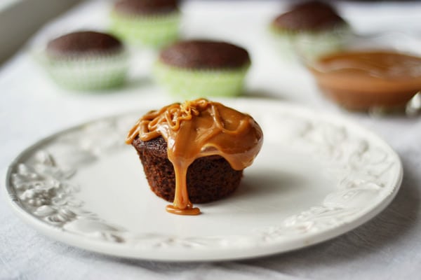 A chocolate cupcake topped with thick caramel sauce sits on a white decorative plate. Nearby, date muffins and a bowl of caramel sauce are blurred in the background.