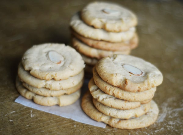 Three stacks of round orange butter cookies sit on a piece of parchment paper on a brown surface. Each golden cookie has a sliced almond on top and looks slightly crisp on the edges.