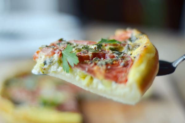 A close-up of a slice of vegetable quiche topped with tomato, herbs, and parsley, being lifted from the whole quiche—perfect alongside Chicken with Sautéed Pasta for a delightful meal.