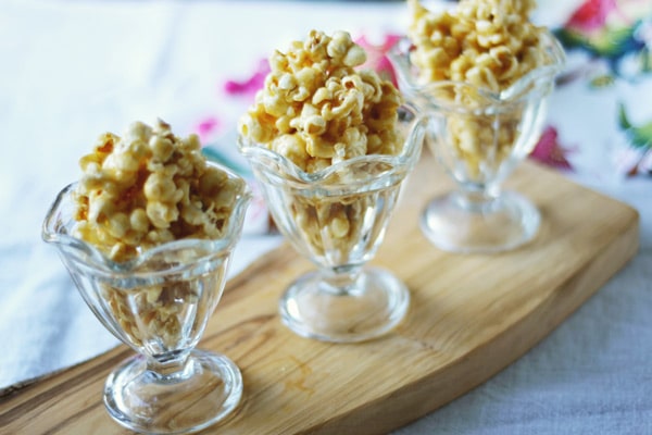 Three glass dessert cups filled with toffee popcorn are arranged in a row on a wooden serving board, with a colorful, floral background slightly out of focus.