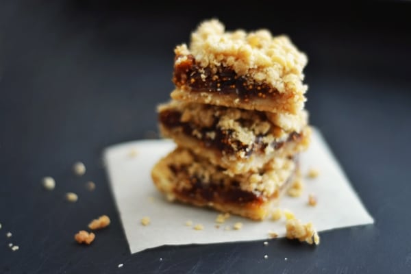 Three Fig Bar Crumble treats are stacked on a small piece of parchment paper, with a few crumbs scattered around, set against a dark background.