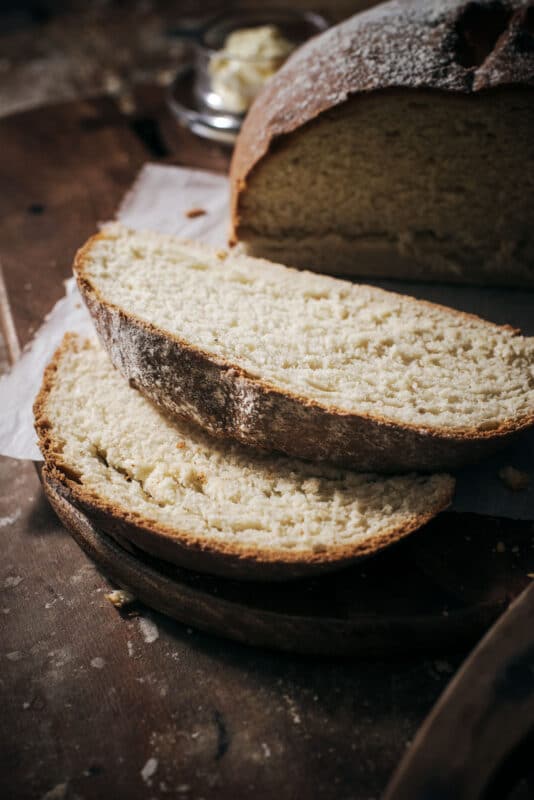 Two thick slices of rustic, homemade bread rest on a wooden board, showcasing the results of a classic White Bread Recipe. The golden-brown crust and soft, airy interior make it irresistible, with butter and the remaining loaf blurred in the background.