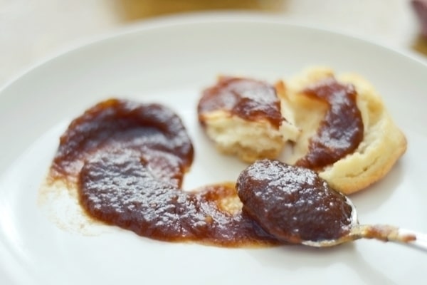 A plate with a biscuit broken in half, generously spread with rich apple butter, and a spoon resting beside it, also covered in the sweet apple butter.