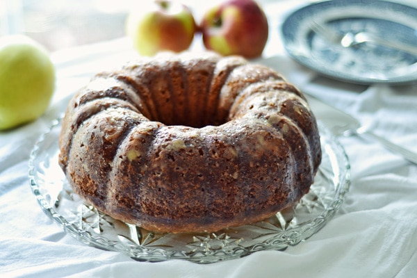 An apple pound cake, beautifully glazed, sits on a glass platter, with fresh apples and blue patterned plates in the background atop a white tablecloth.