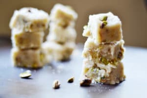 Three stacks of white cardamom fudge, classic Indian sweets likely made with milk and pistachios, are placed on a dark surface with scattered pistachio pieces. The focus is on the front stack; the background is softly blurred.