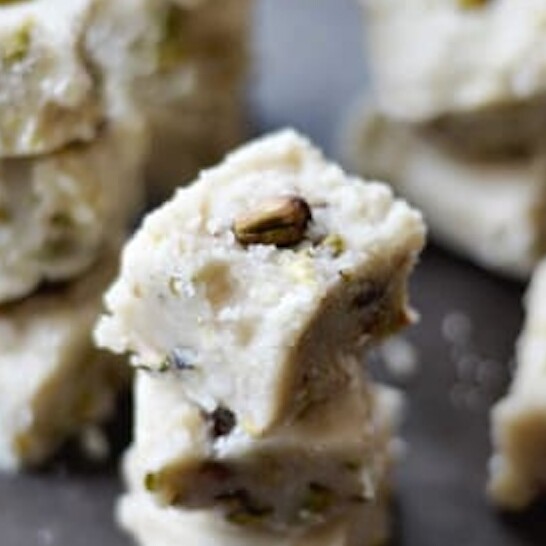 A close-up of stacked pieces of white Indian cardamom fudge, likely made of milk and sprinkled with pistachios, placed on a dark surface. Some pieces are blurred in the background.