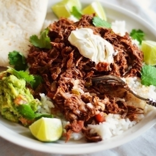 A plate of shredded beef, inspired by Chilli Con Carne, served with white rice, guacamole, lime wedges, cilantro, a dollop of sour cream, and a tortilla on the side. A fork rests on the plate.