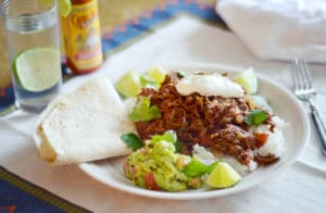 A plate of Chilli Con Carne shredded meat on rice, topped with sour cream, served with guacamole, lime wedges, fresh cilantro, and a tortilla. A glass of water with lime and a bottle of hot sauce are nearby.