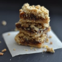 A stack of three crumbly, golden brown Fig Bar Crumble dessert bars with a dark fruit filling sits on parchment paper, surrounded by scattered crumbs.