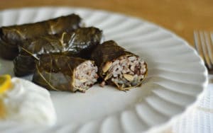 Three Stuffed Grape Leaves filled with rice and pine nuts sit on a white plate, served with a dollop of yogurt. One roll is cut open to reveal the savory filling, while a fork rests on the right.