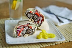 A Mediterranean Wrap cut in half, filled with rice, vegetables, and feta cheese, is served on a square white plate with yellow pepper slices. A glass of water with lemon and a napkin with forks are in the background.