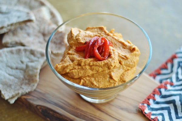 A glass bowl of creamy Roasted Red Pepper Hummus, topped with sliced roasted red peppers, sits on a wooden board next to pita bread and a chevron-patterned napkin.