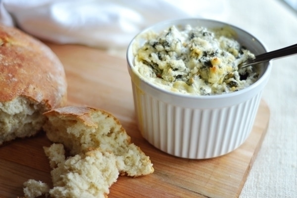 A white ramekin filled with creamy Spinach Artichoke Dip sits on a wooden board next to torn pieces of rustic bread. A spoon rests in the dip, and a white cloth is in the background.