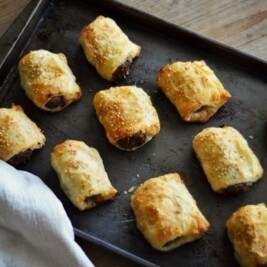 A baking tray with ten golden-brown Sausage Rolls, some sprinkled with sesame seeds, sits on a wooden surface next to a white cloth.