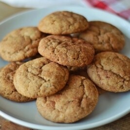 A white plate holds a pile of golden-brown snickerdoodles with a slightly cracked surface. A red and white checked cloth is partially visible in the background on a wooden table.