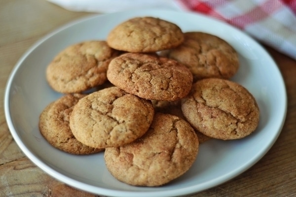 A white plate holds a pile of golden-brown snickerdoodles with a slightly cracked surface. A red and white checked cloth is partially visible in the background on a wooden table.