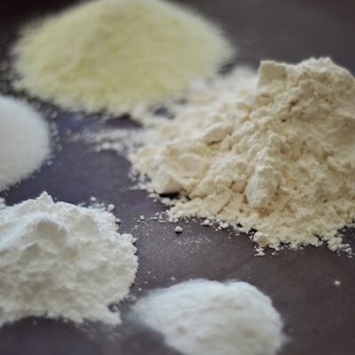 Five small piles of various white and off-white powders, likely common baking ingredients such as flour, sugar, and baking powder—or even an All Purpose Baking Mix—are arranged on a dark surface.