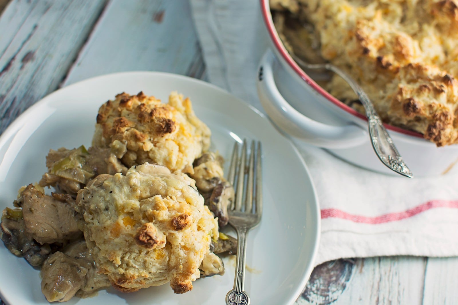 A white plate holds a serving of biscuit-topped chicken and vegetable cobbler casserole, with two forks beside it. In the background, a baking dish with more cobbler casserole sits on a white cloth.