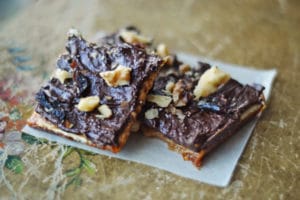 Two pieces of Chocolate Toffee Brittle, coated in rich chocolate and topped with chopped nuts, rest on a small piece of parchment paper on a textured surface.
