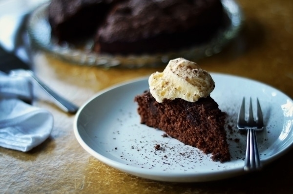 A slice of rich Chocolate Fudge Cake topped with a scoop of vanilla ice cream sits on a white plate with a fork, while the rest of the cake is blurred in the background.