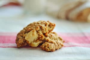 A close-up of Oatmeal Apple Cookies, with visible apple chunks, broken in half and resting on a white cloth with a pink stripe. The softly blurred background adds warmth to the inviting scene.