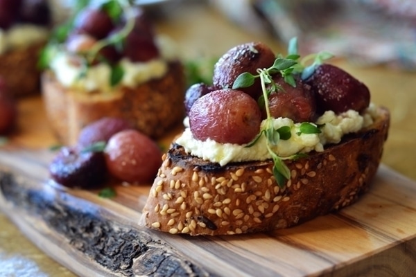 A slice of seeded bread topped with whipped cheese, roasted grapes, and fresh herbs, this Roasted Grapes Crostini is served on a rustic wooden board.