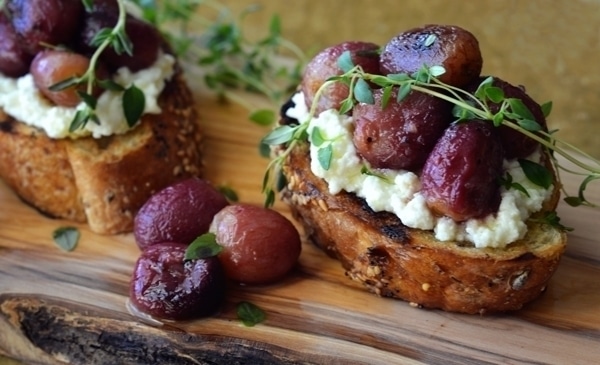 Two slices of toasted bread topped with creamy ricotta cheese, roasted grapes, and fresh thyme make this Roasted Grapes Crostini a delightful treat, served on a wooden board with extra grapes and herbs as garnish.