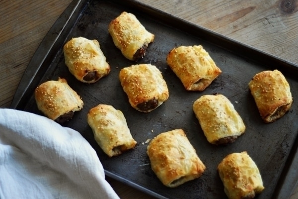 A baking tray with ten golden-brown Sausage Rolls, some sprinkled with sesame seeds, sits on a wooden surface next to a white cloth.