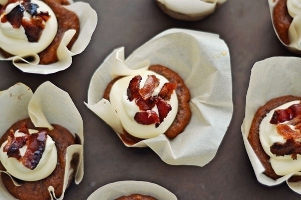 Sweet Potato Muffins with Bacon and Maple Buttercream in parchment wrappers, topped with swirls of white frosting and crispy bacon pieces, arranged on a dark surface.