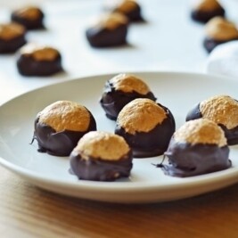 A white plate holds several Buckeyes: round peanut butter balls partially dipped in chocolate, leaving the tops exposed. More Buckeye candies are visible on a tray in the background.