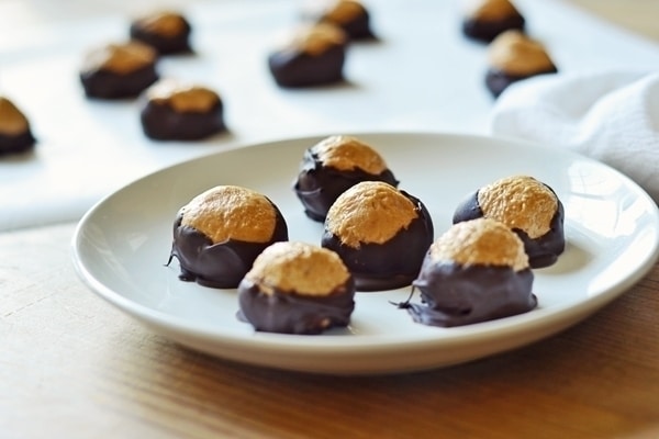 A white plate holds several Buckeyes: round peanut butter balls partially dipped in chocolate, leaving the tops exposed. More Buckeye candies are visible on a tray in the background.