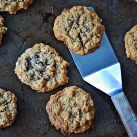 A metal spatula lifts a freshly baked Cowboy Cookie, loaded with oatmeal and chocolate chips, from a metal baking sheet surrounded by several more cookies.