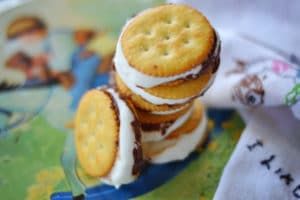 Three Ritzy Smores sandwich cookies made with round crackers and a creamy white filling are stacked on a colorful plate, with a white napkin featuring small illustrations on the side.