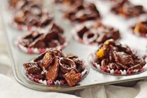 Close-up of a muffin tray filled with paper liners, each holding clusters of Easy Chocolate Covered Snack Mix, including pretzels and cereal pieces. The glossy treats are arranged in neat portions for a simple, delicious snack.