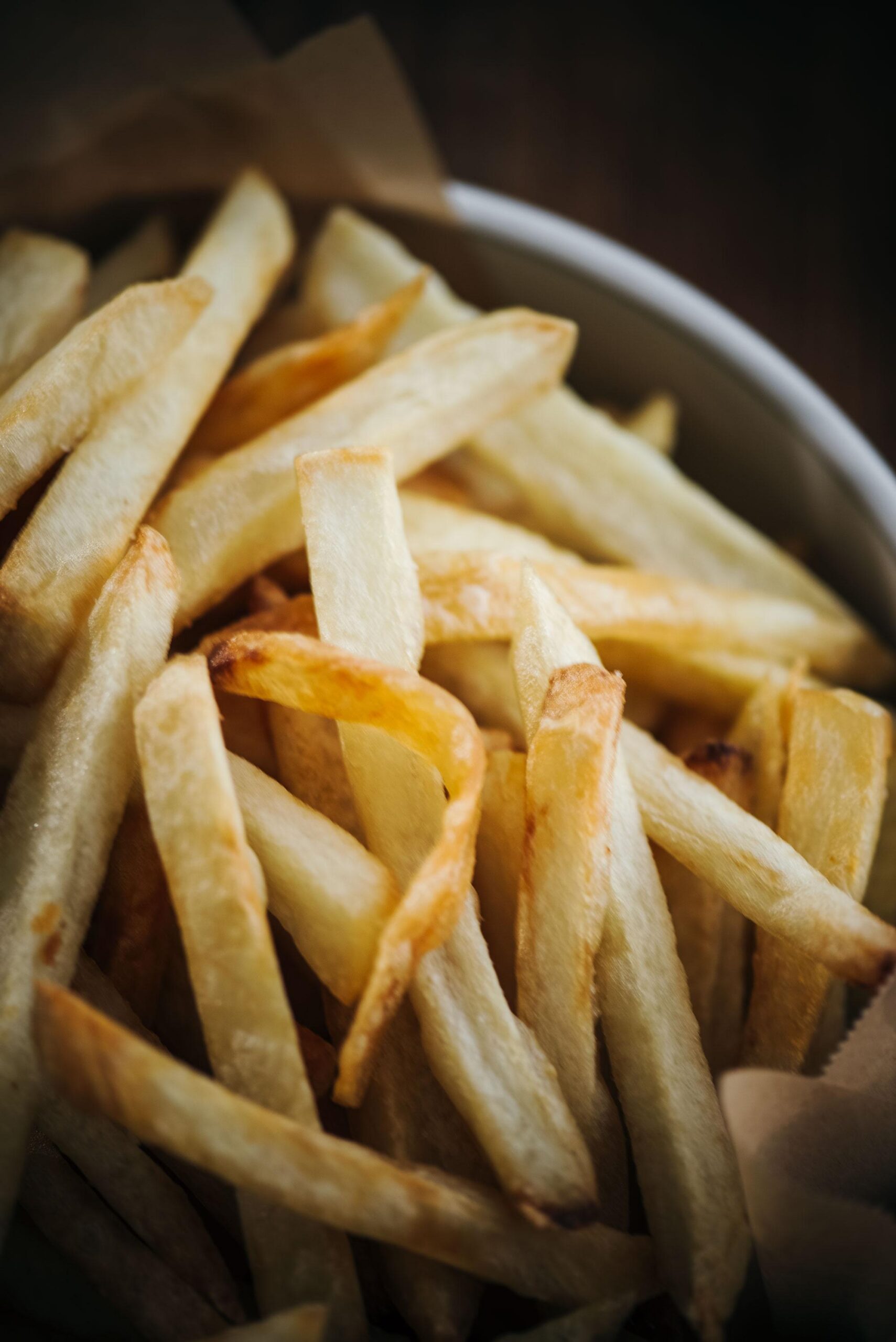 A close-up of golden, crispy French Fries piled in a bowl, showcasing their slightly uneven, hand-cut shapes and perfectly browned edges.