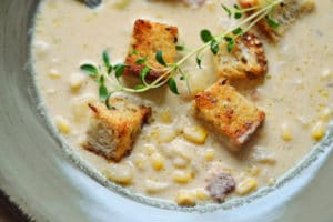 A bowl of creamy Corn Chowder with Smokey Bacon, white beans, and corn, topped with thyme scented croutons and garnished with fresh sprigs of thyme.