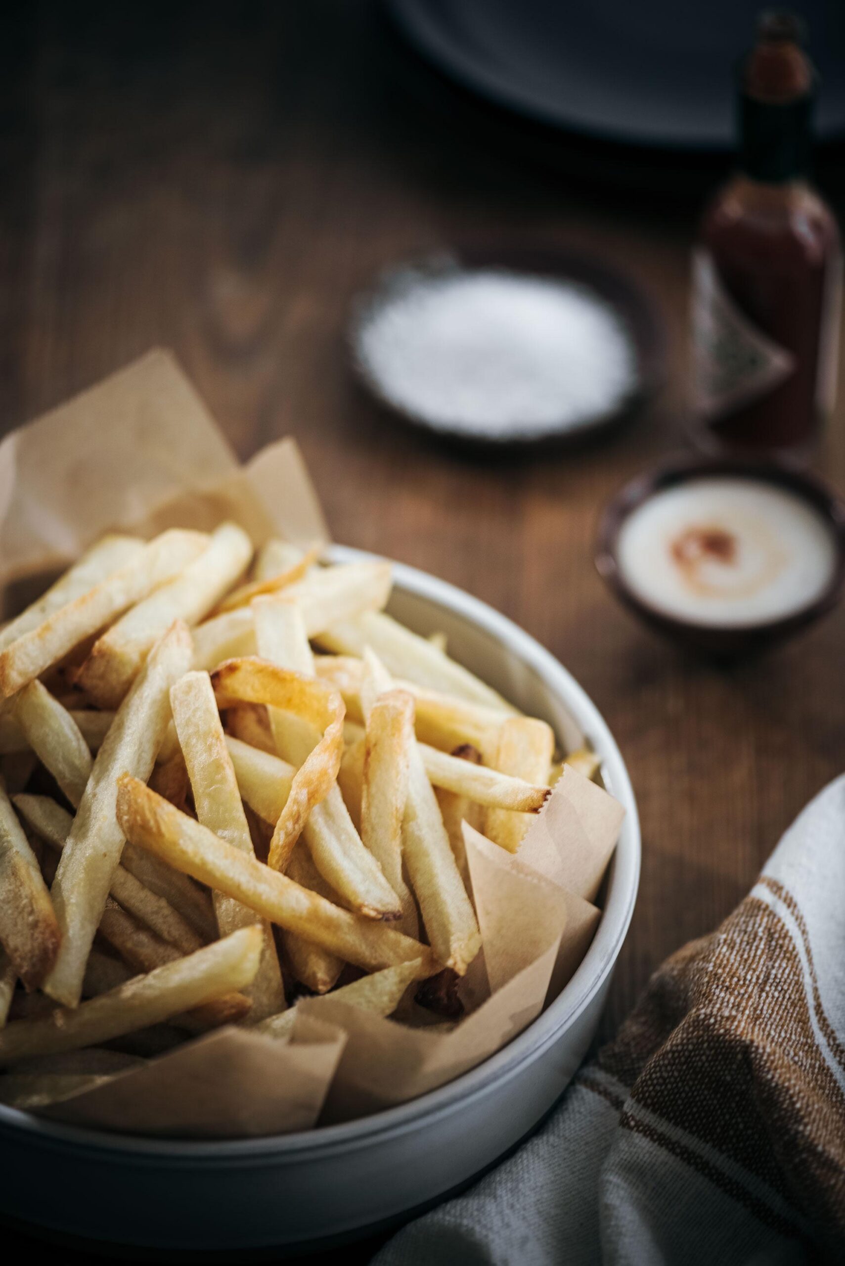 A bowl of crispy French fries sits on brown parchment paper atop a wooden table, accompanied by a small dish of salt, a bowl of dipping sauce, a bottle of hot sauce, and a brown-and-white striped napkin.