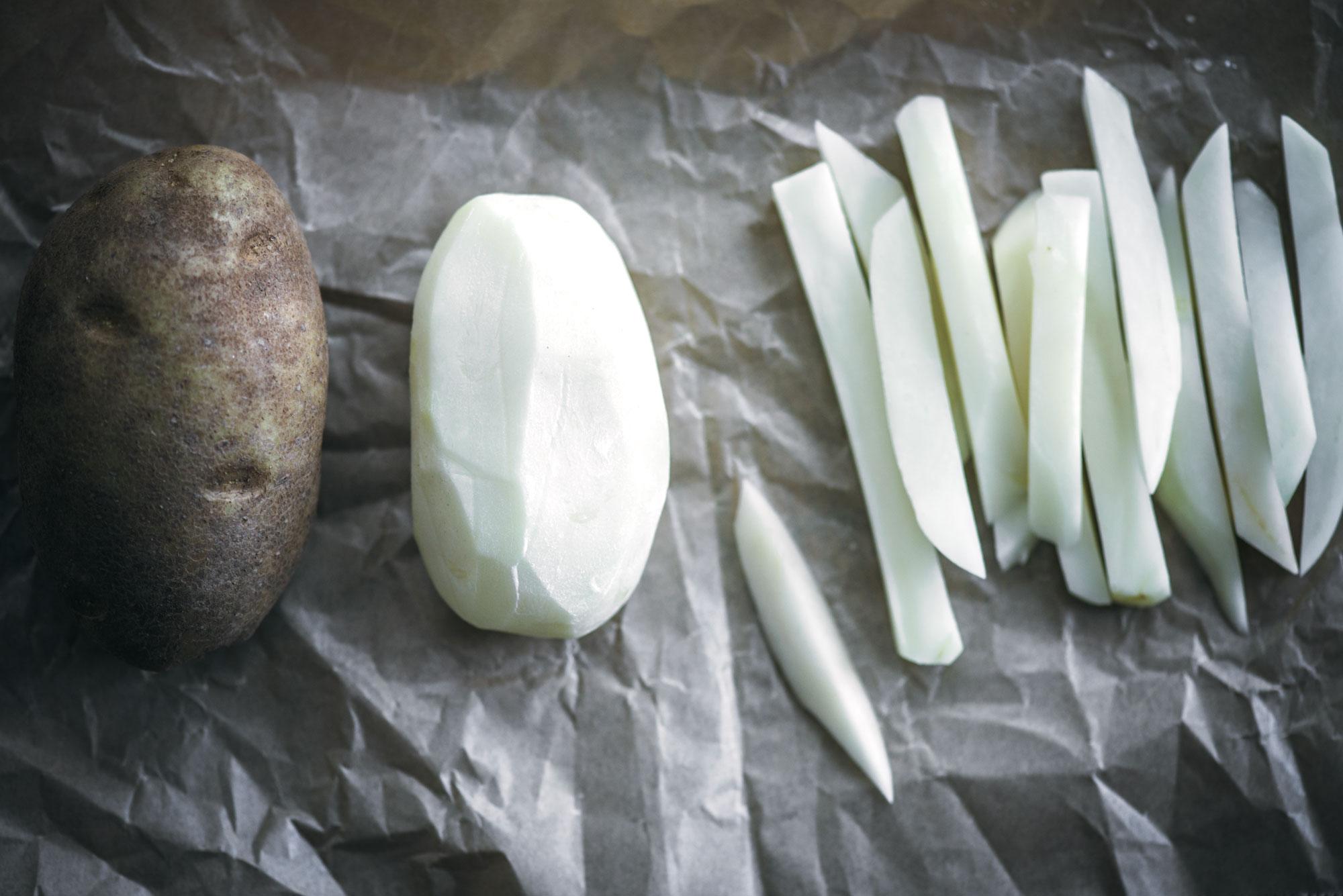A whole unpeeled potato, a peeled potato, and several raw strips—perfect for making crispy French fries—are arranged side by side on a crumpled sheet of parchment paper.