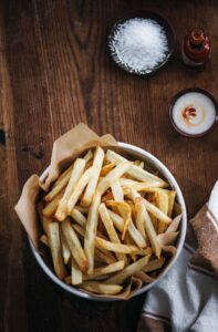A bowl of crispy French fries sits on a wooden table, lined with brown parchment paper. Nearby are a small bowl of salt, a dish of dipping sauce, and a bottle of hot sauce, with a brown and white cloth partially visible.