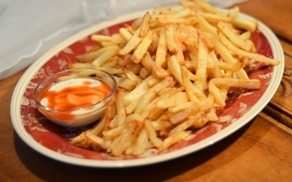 A plate of golden, crispy French Fries is served with a small bowl of creamy dipping sauce topped with a swirl of reddish-orange sauce, all set on a wooden table.