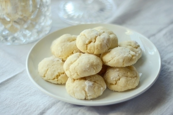 A white plate with a pile of round, pale Key Lime Cookies sits on a white cloth. The cookies have a slightly cracked surface and a soft, crumbly appearance, hinting at their zesty citrus flavor.